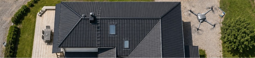 Flygfoto uppifrån av ett villatak, med en drönare i bild över grusytan intill huset